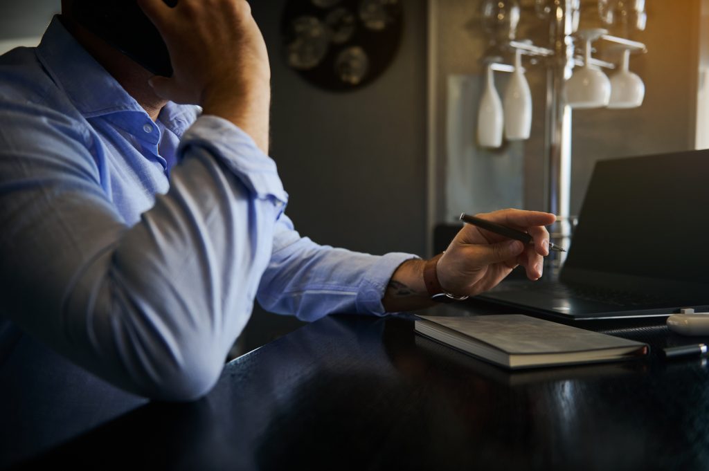 Focus on the hand holding a pen of a busy unrecognizable businessman, freelancer talking on mobile phone while working remotely on laptop at home office